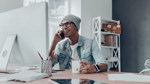 A young man sits at a desk in front of a desktop computer with a cell phone held up to his ear.